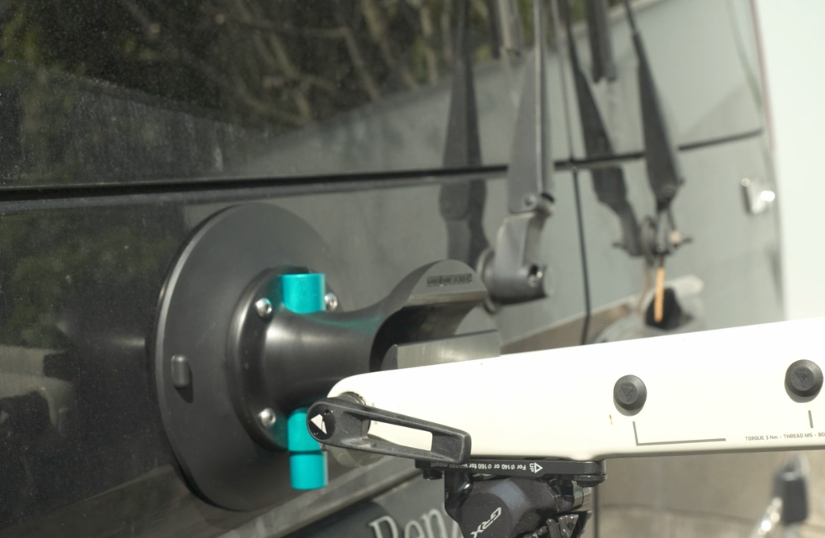 Eine Nahaufnahme des Saugnapfhalters Fahrrad mit Steckachsenadapter, der an einem dunklen Fahrzeug befestigt ist, mit einem Teil eines weißen Fahrrads auf dem Gepäckträger und Scheibenwischern im Hintergrund.