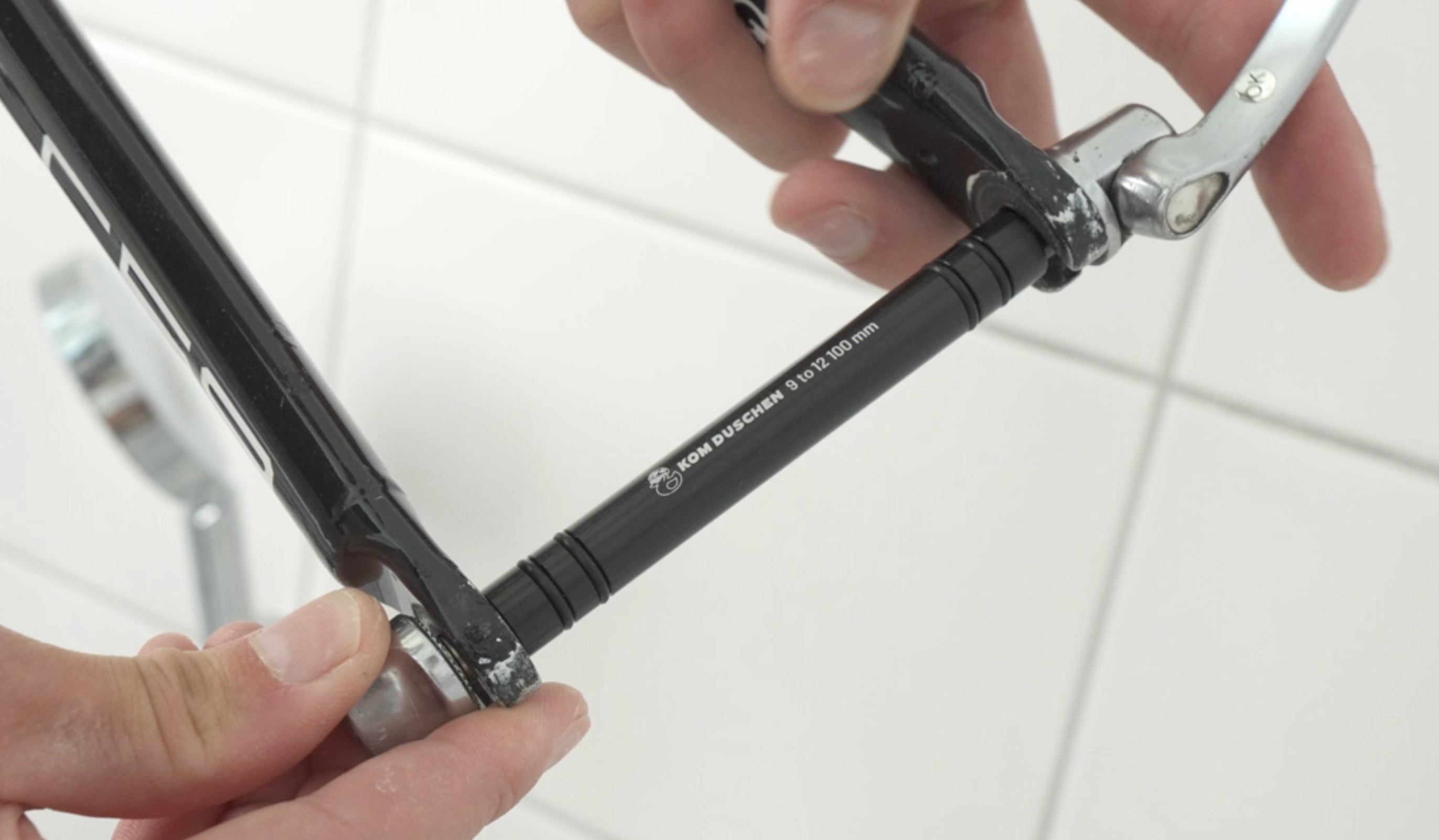 Nahaufnahme von Händen, die einen Schnellspanner-Adapter an einem Fahrradrahmen montieren, mit einer weiß gekachelten Wand im Hintergrund.