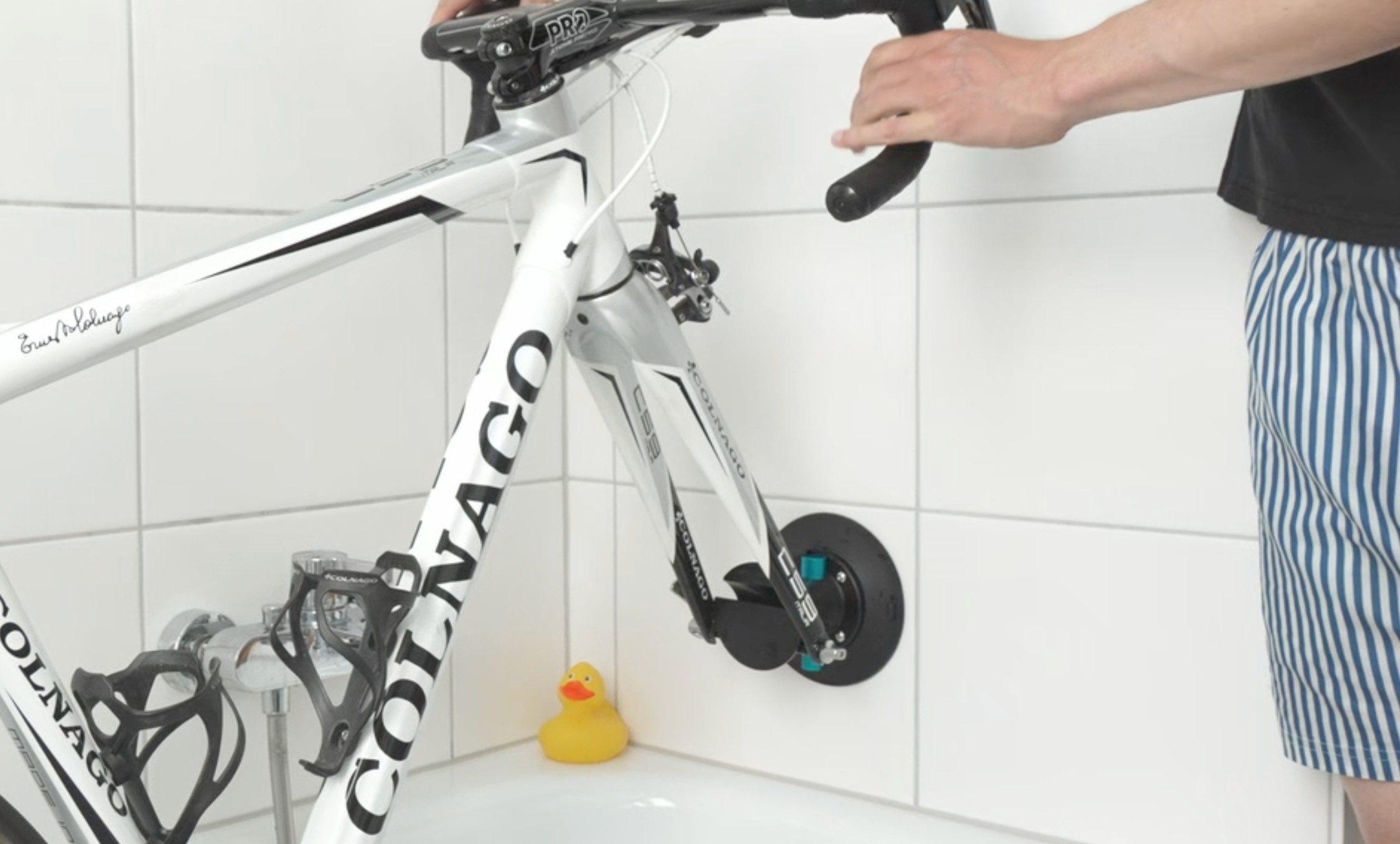 Eine Person in gestreiften Shorts hält den Lenker eines weißen Colnago-Fahrrads, das in einer Badewanne mit einer Gummi-Ente in der Nähe abgestellt ist. Das Fahrrad ist mit dem Saugnapfhalter Fahrrad an einer gekachelten Wand befestigt.