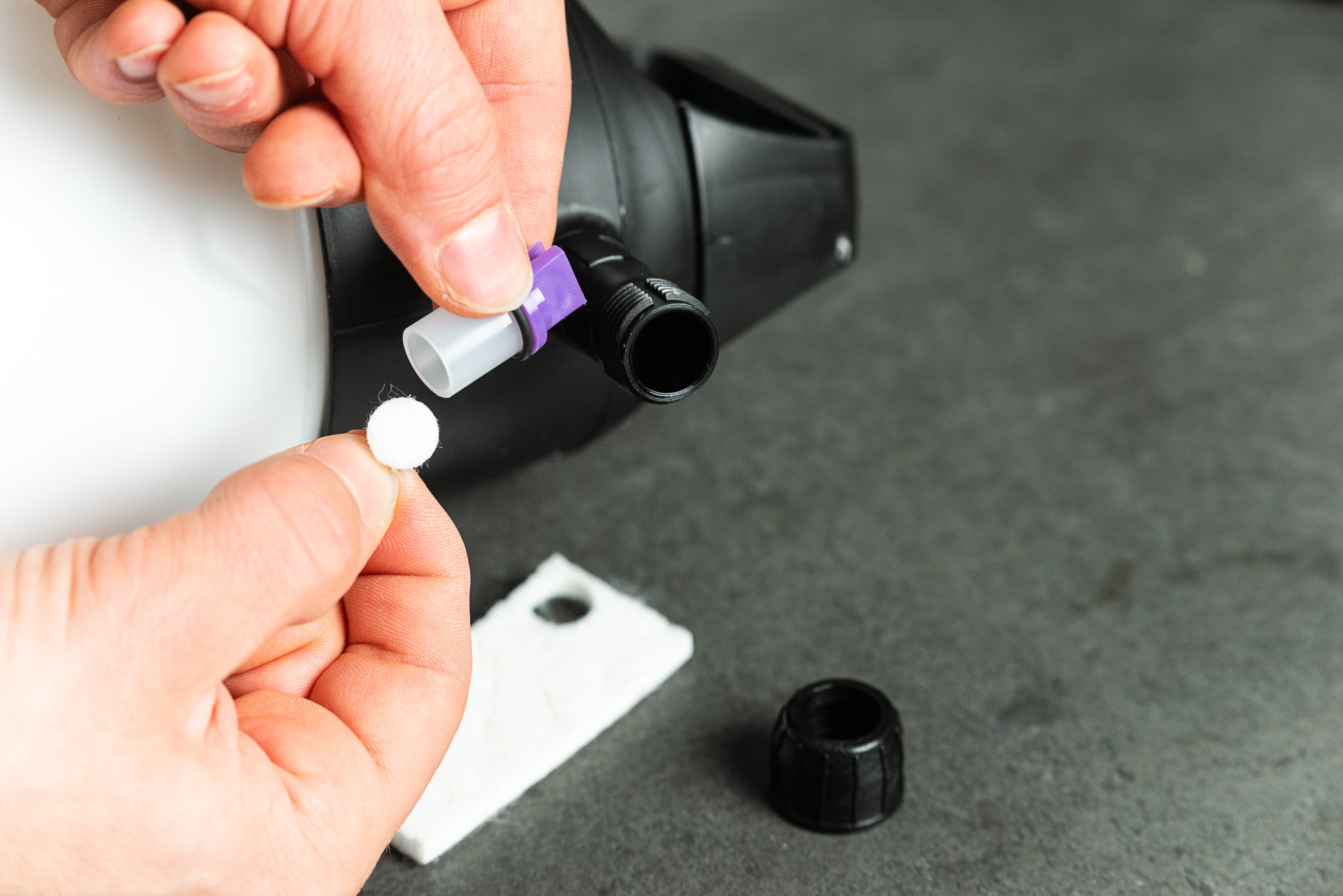 Die Hände stecken eine weiße Tablette in die Düse des IK FOAM Pro 2 - Pumpsprühers, die Kappe ist abgenommen und ein weißes Filterpad liegt in der Nähe auf einer grauen Fläche, bereit für eine effiziente Fahrradreinigung mit dem Schaumsystem.