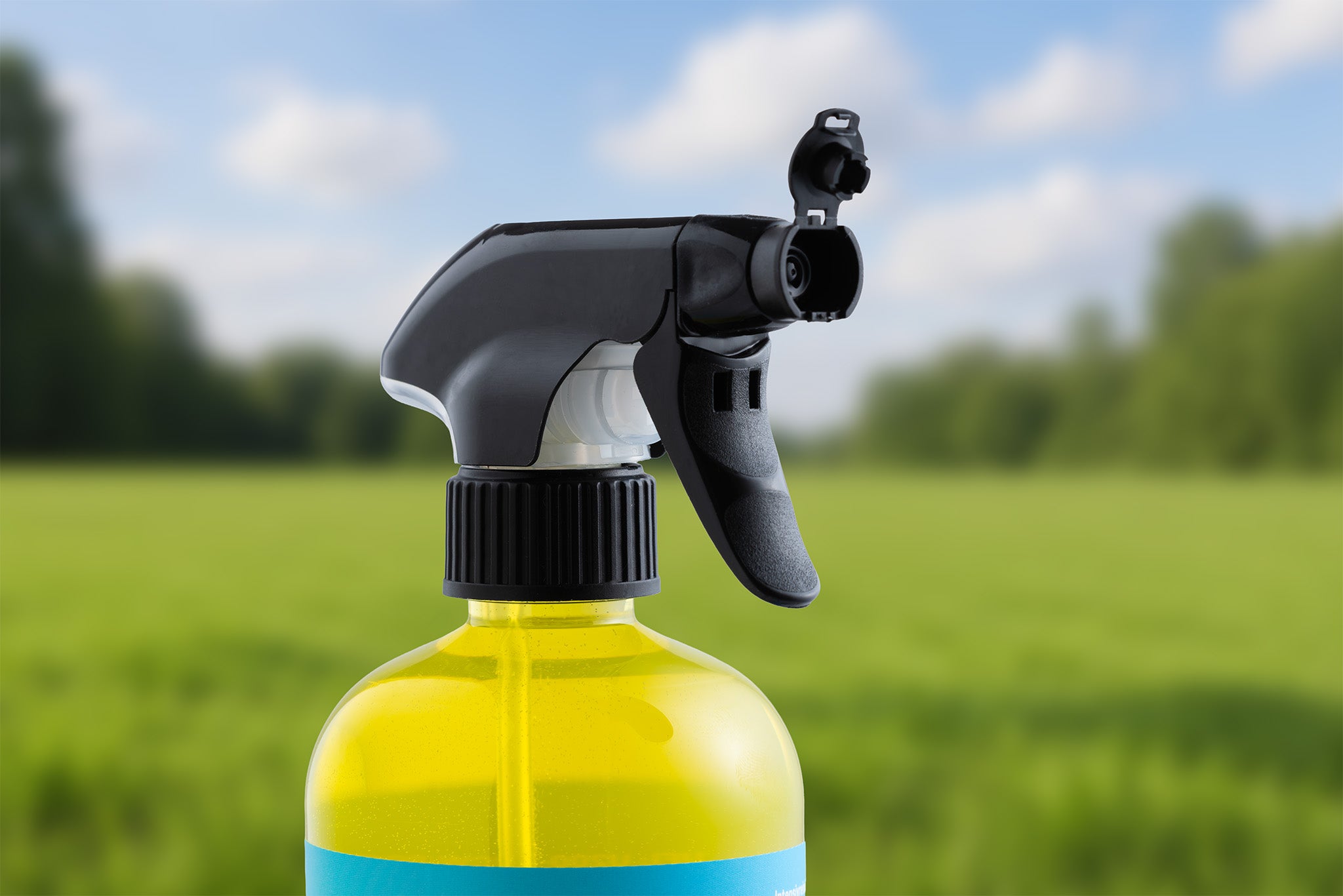 Eine Nahaufnahme einer Intensivreiniger - Ready to Use - Sprühflasche mit gelbem Reiniger, schwarzem Sprühkopf und gelartiger Formulierung vor einem unscharfen Hintergrund aus grünem Gras und Bäumen unter blauem Himmel.