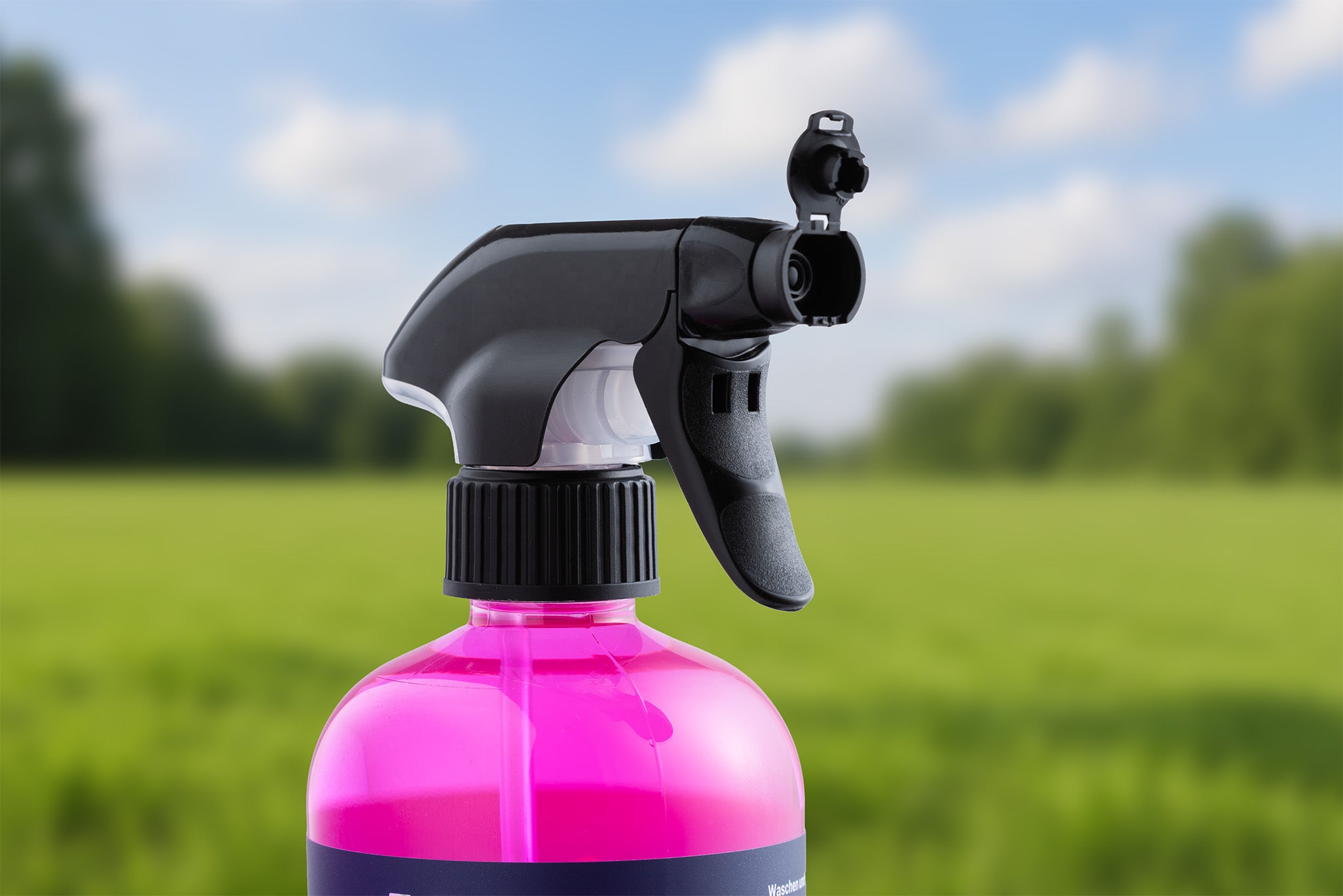 Nahaufnahme einer Sprühflasche mit leuchtend pinkfarbener Flüssigkeit "Waschen und Versiegeln - Ready to Use" und einer schwarzen Sprühpistole vor einem unscharfen grünen und blauen Hintergrund im Freien, wobei der Schwerpunkt auf dem Lackschutz liegt.
