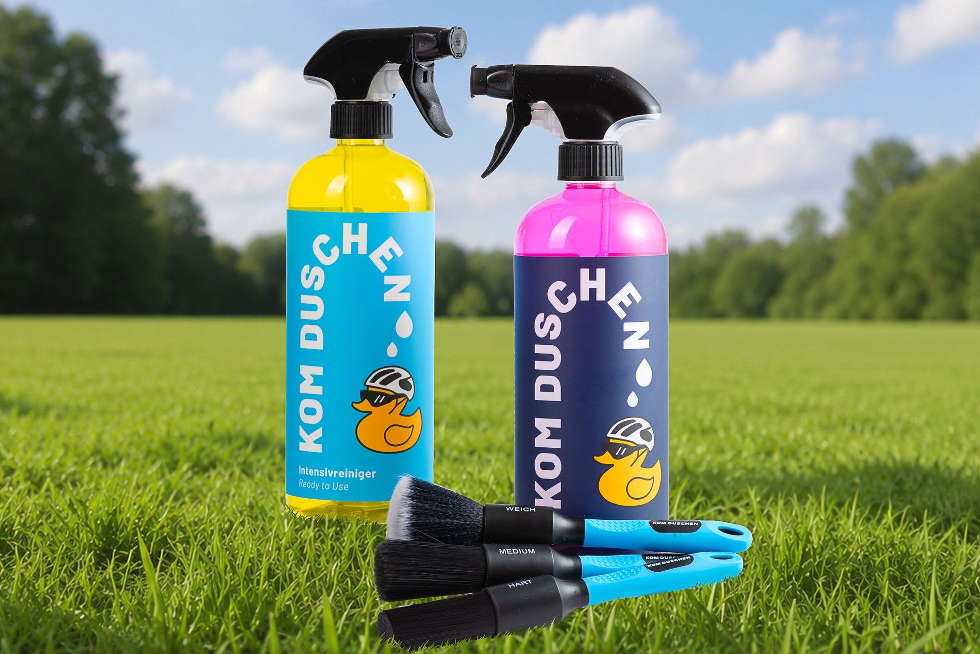 Zwei Sprühflaschen aus dem Essential Plus - Set, mit einem Entenlogo, stehen auf Gras neben einem Satz von drei Bürsten. Ideal zum Reinigen und Versiegeln, mit Bäumen und blauem Himmel sanft verschwommen im Hintergrund.