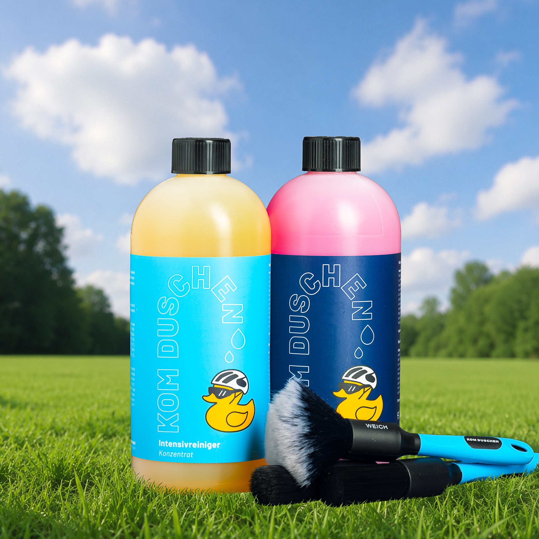 Zwei Flaschen „Essential Plus Konzentrate“ – Reinigungsset, eine gelbe und eine rosa, stehen auf einer Wiese, davor liegen zwei Bürsten. Auf den Flaschen ist eine gelbe Ente mit Sonnenbrille abgebildet. Im Hintergrund sind ein strahlend blauer Himmel und Bäume zu sehen.