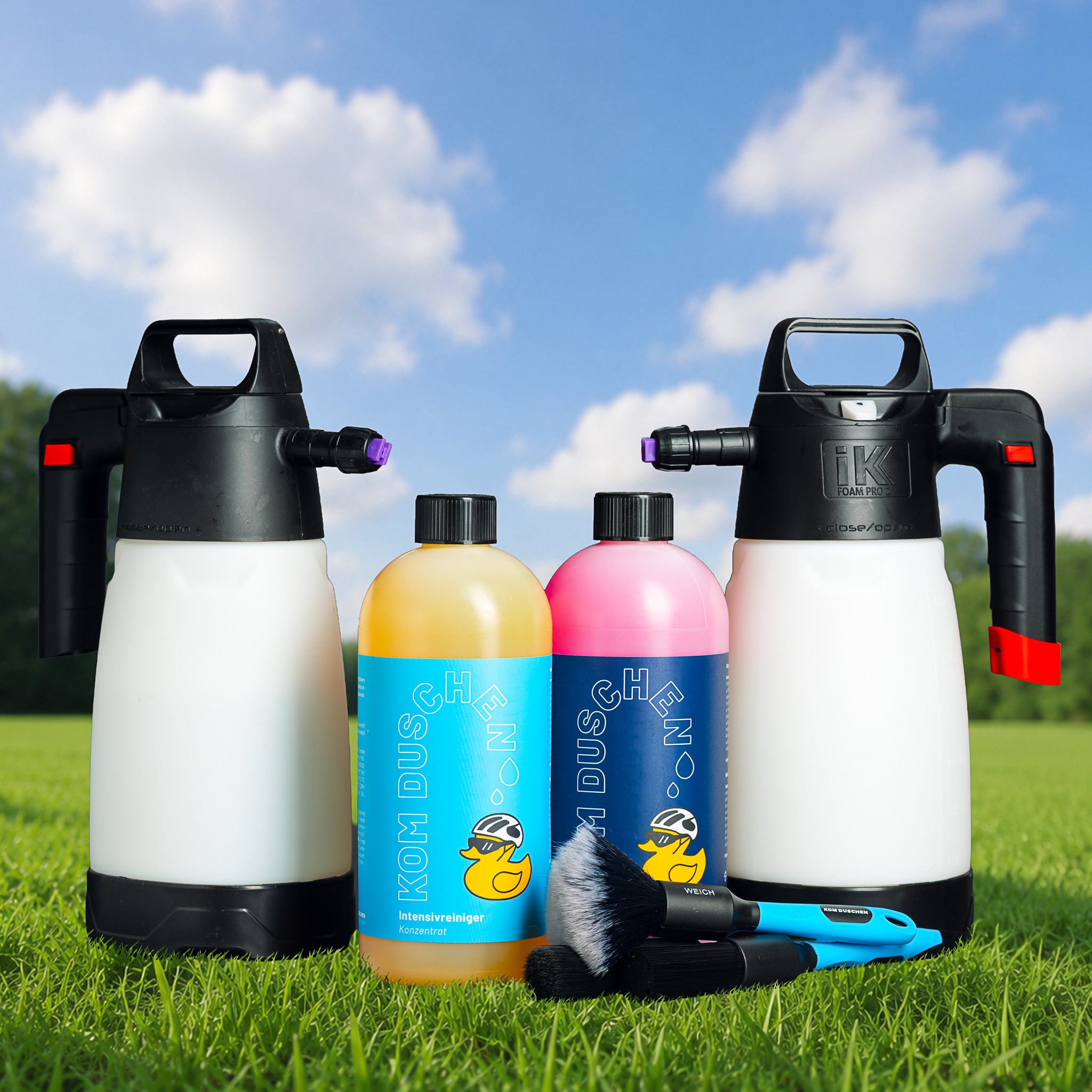 Das „Profi-Konzentrat“-Set, bestehend aus zwei großen Sprühflaschen, einer gelben und einer rosa Reinigungsflasche sowie zwei Reinigungsbürsten, wird vor dem Hintergrund von grünem Gras, Bäumen und einem blauen Himmel präsentiert.