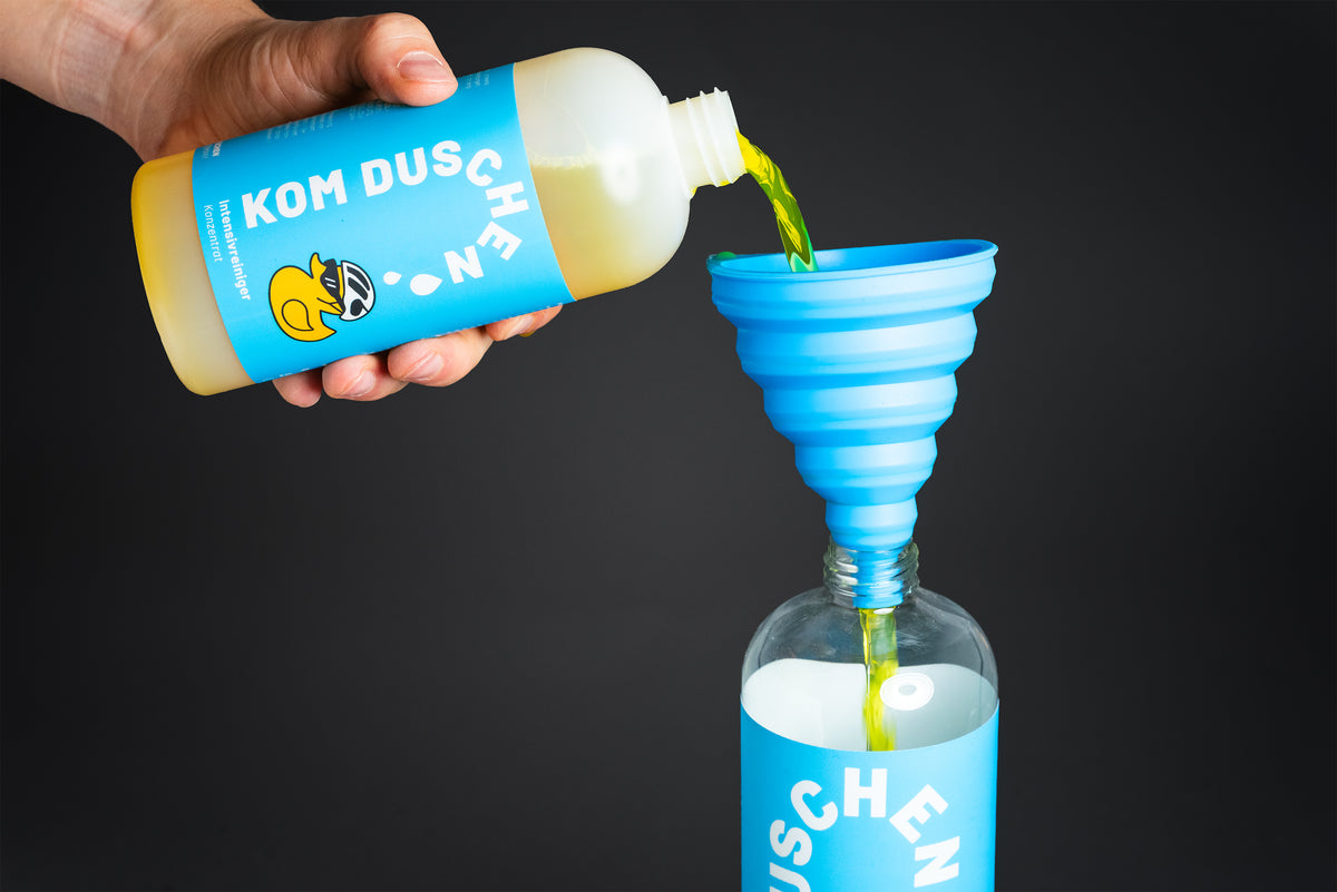 Eine Hand gießt gelbe Flüssigkeit aus einer KOM DUSCHEN-Flasche in einen blauen Silikontrichter, der in einer weiteren, dazu passenden Flasche – ideal für Reinigungslösungen – vor einem schwarzen Hintergrund steht.